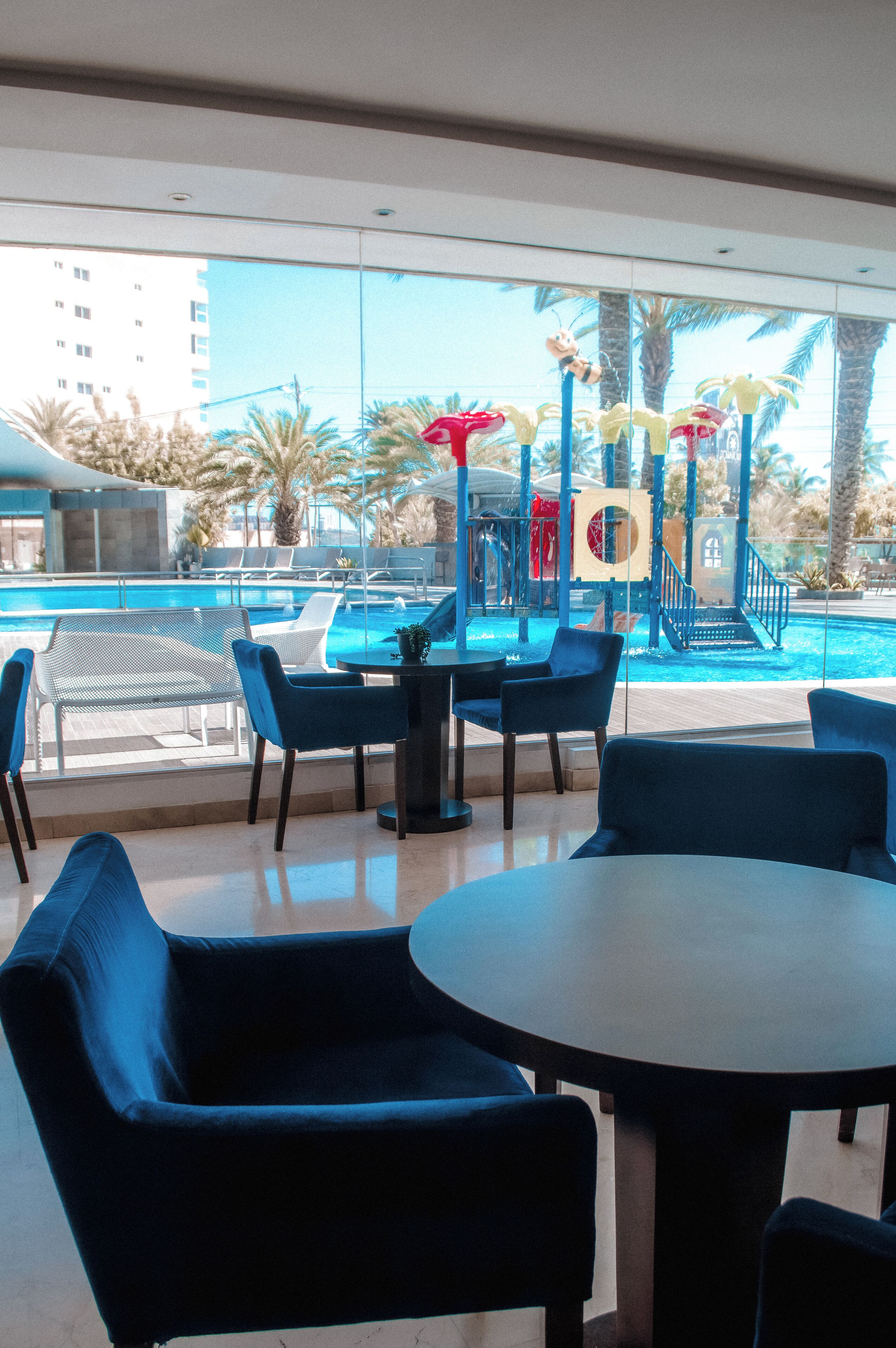 A poolside area with blue chairs and a table, overlooking a swimming pool and palm trees.