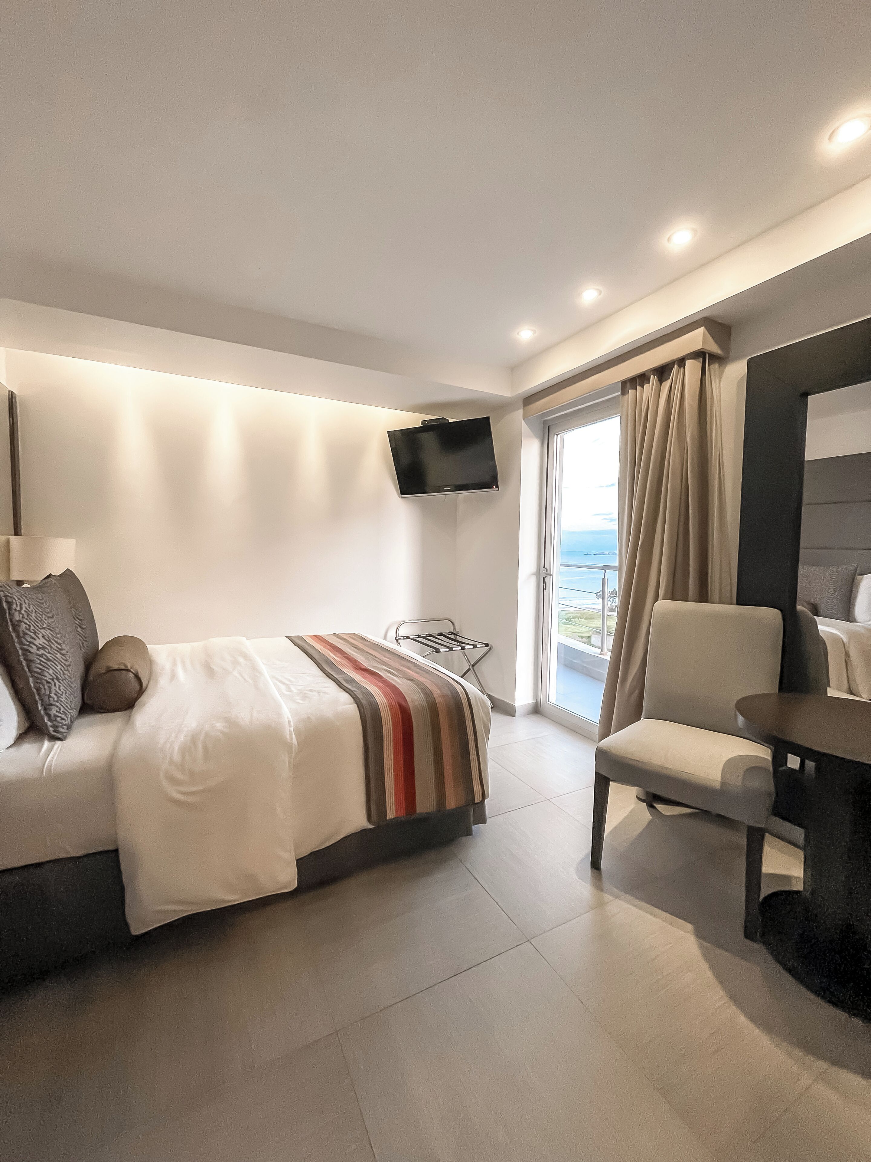 A modern hotel room with a bed, a chair, a TV mounted on the wall, and a view of the outdoors.