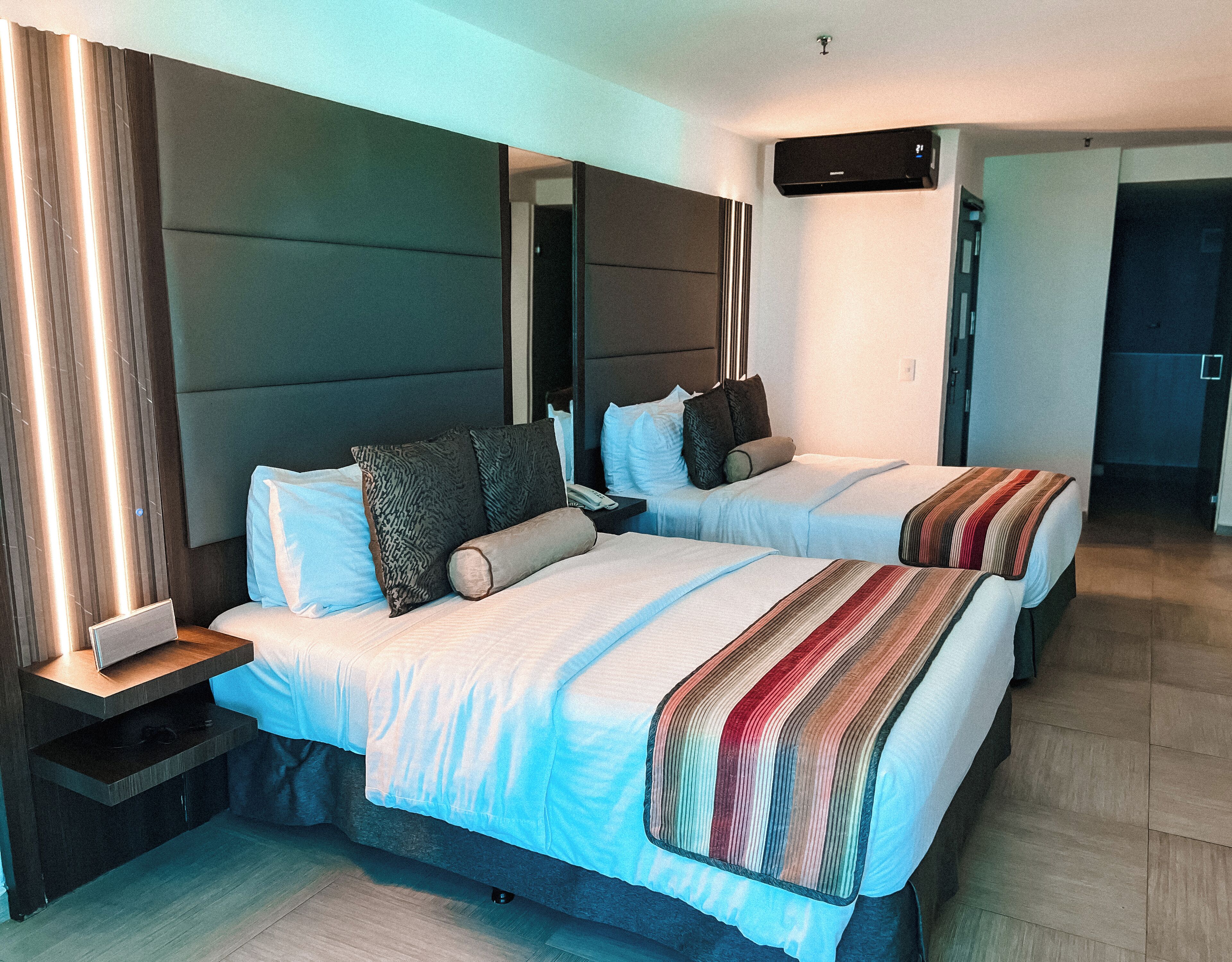 A hotel room with two beds, a bedside table, a nightstand, a mirror, and a built-in air conditioning unit.