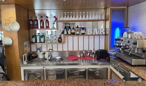 A bar area with a stainless steel countertop, a coffee machine, and shelves with bottles and glasses. - Hotel Bel Air (Rimini)