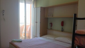 A single bed with a striped bedspread, a wooden headboard, and a small bedside table. There is a white chair outside the door, and a red telephone on a shelf above the bed. - Hotel Bel Air (Rimini)