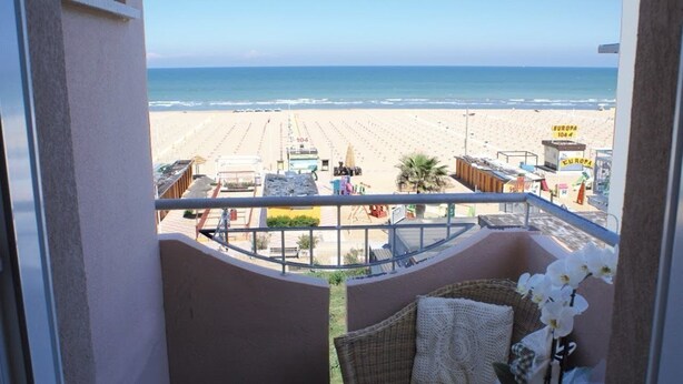 A balcony with a wicker chair and a small table, overlooking a beach with a sign reading "EUROPA 104". - Hotel Bel Air (Rimini)