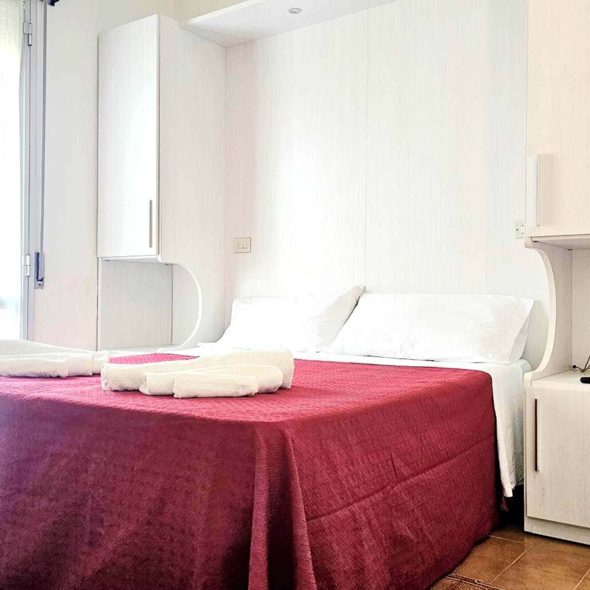 A hotel room with a bed, white walls, a red bedspread, and a white nightstand.