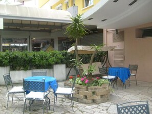 An outdoor patio area with tables and chairs, a small garden with flowers, and a building in the background. - Hotel Bel Air (Rimini)