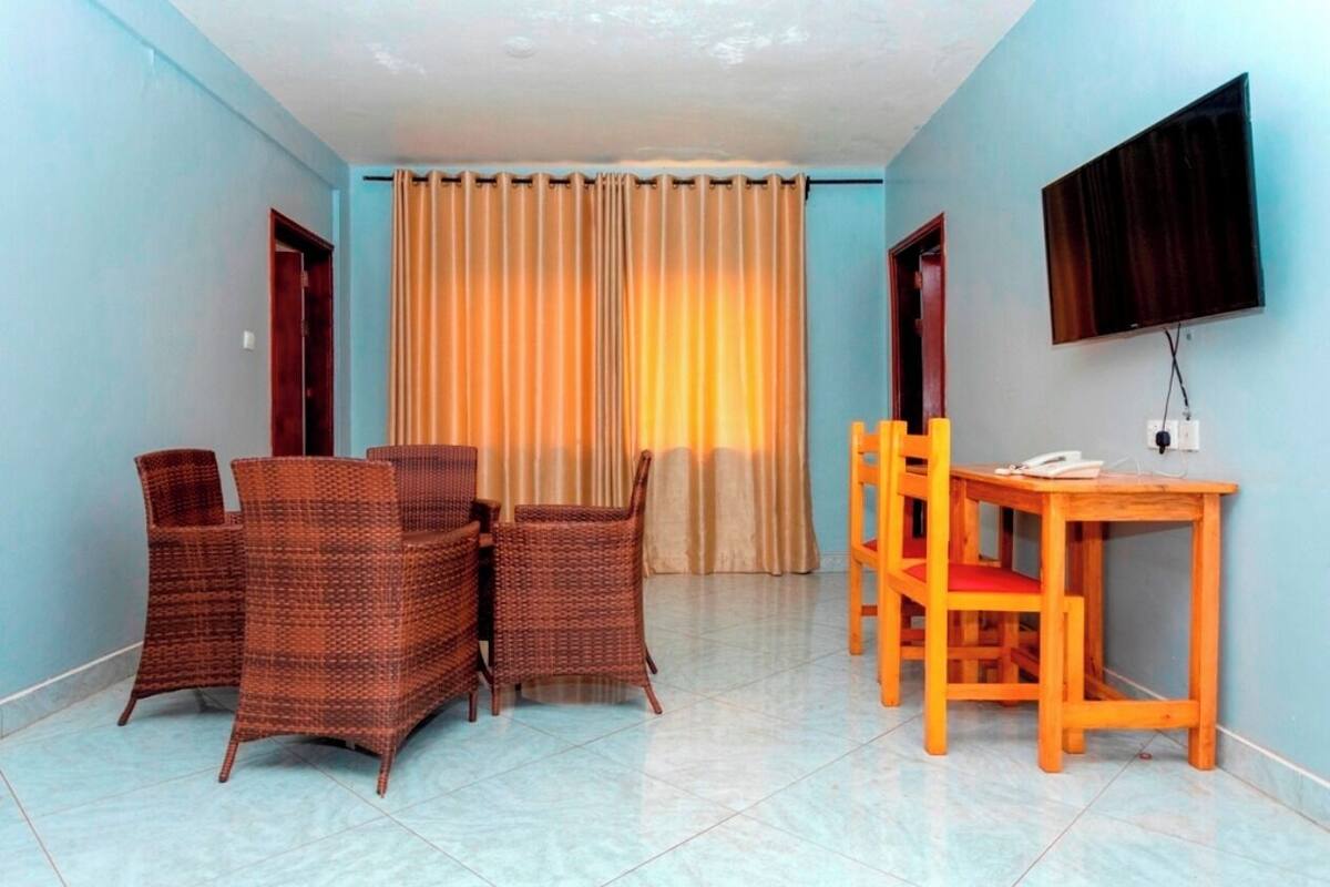 A room with a dining table and chairs, a TV mounted on the wall, and a window with curtains.