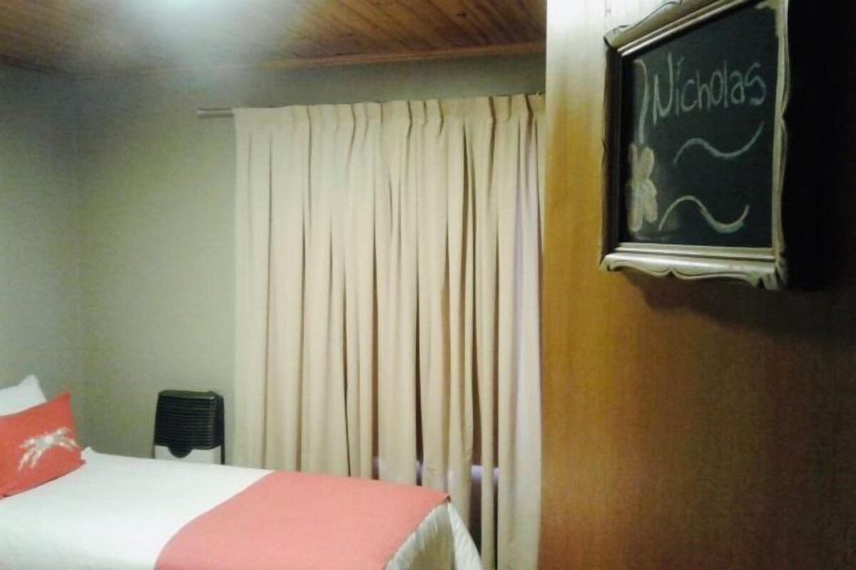A hotel room with a bed, a chair, a radiator, and a chalkboard with the name "Nicholas" written on it.