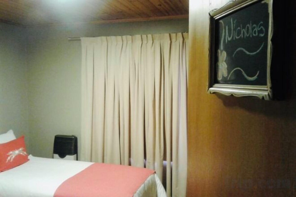 Une chambre d’hôtel avec un lit, une chaise, une fenêtre avec des rideaux et un tableau noir sur lequel est écrit le nom « Nicholas ».