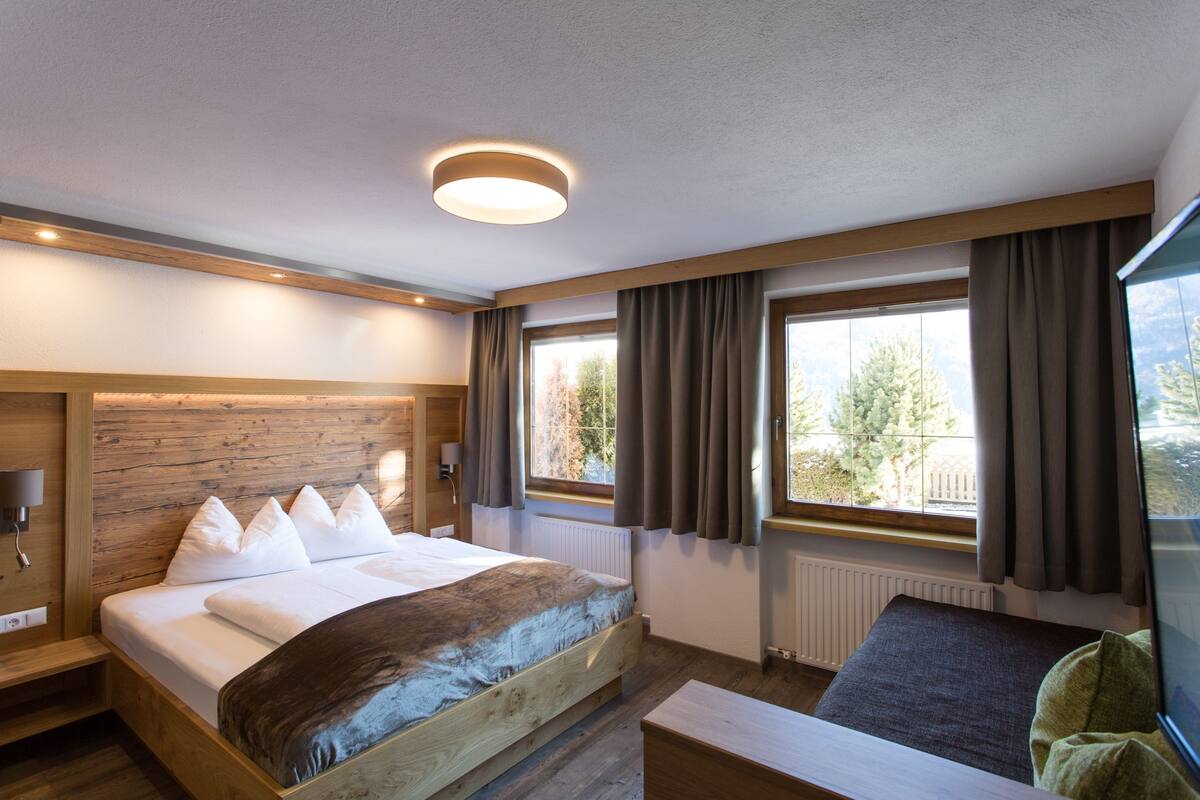 A modern hotel room with a wooden bed, a large window with curtains, a wooden bench, and a ceiling light.