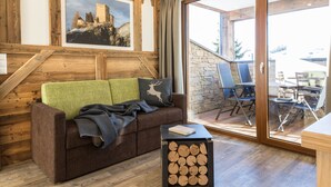 A cozy room with a wooden wall, a green and black sofa, a small table with logs, and a glass door leading to an outdoor seating area.