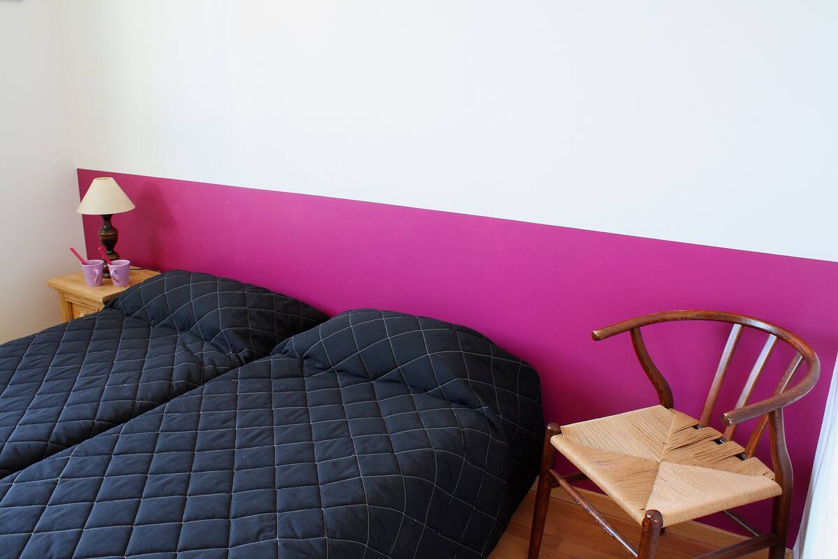 A bed with a quilted cover, a wooden chair, a small table with a lamp, and a pink wall.