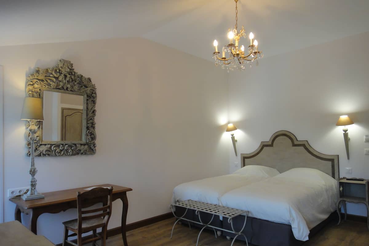 A bedroom with a double bed, a desk, a chair, a small table, a mirror, and a chandelier.