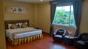 A hotel room with a large bed, two armchairs, a small table, a window with curtains, and a wall-mounted artwork.