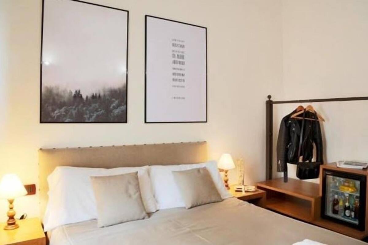 A hotel room with a bed, two framed pictures on the wall, a coat rack with a jacket, and a small table with a lamp and a bottle.