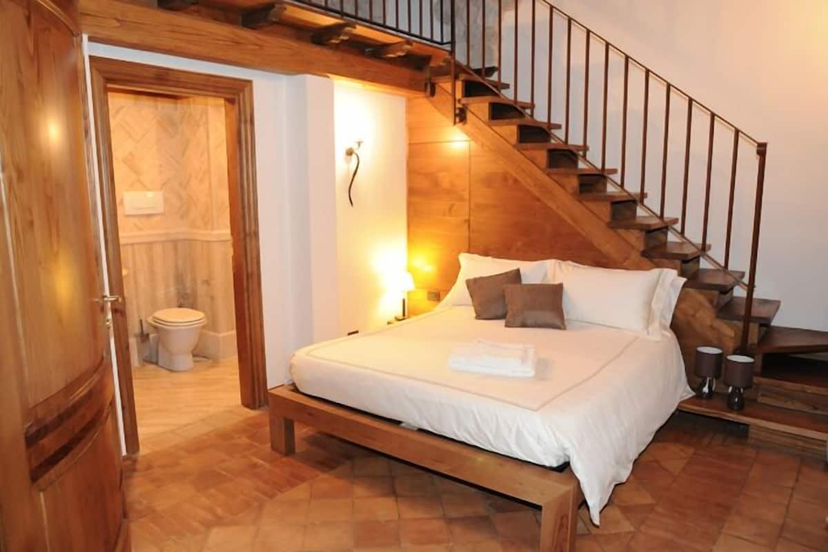 A bedroom with a wooden bed, a nightstand, a lamp, a wooden staircase, and a bathroom visible through an open door.