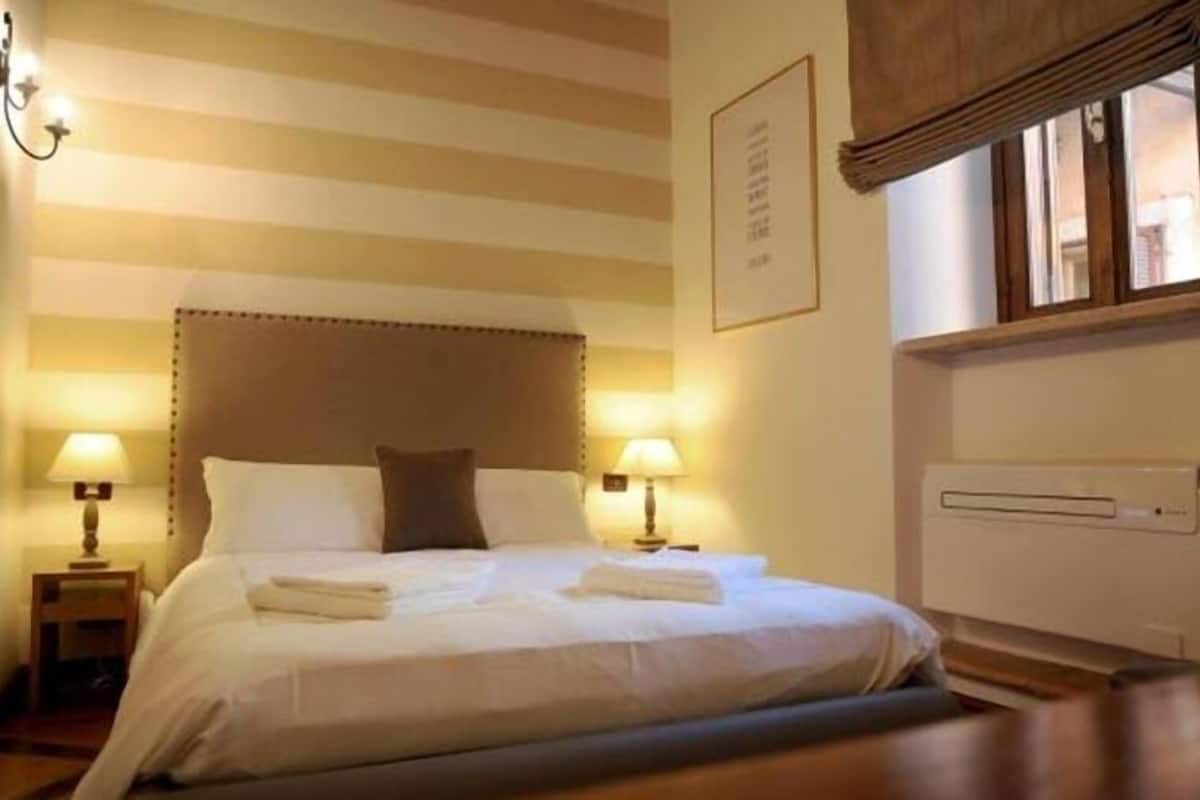 A hotel room with a bed, two bedside lamps, a window with blinds, and a wall-mounted air conditioning unit.