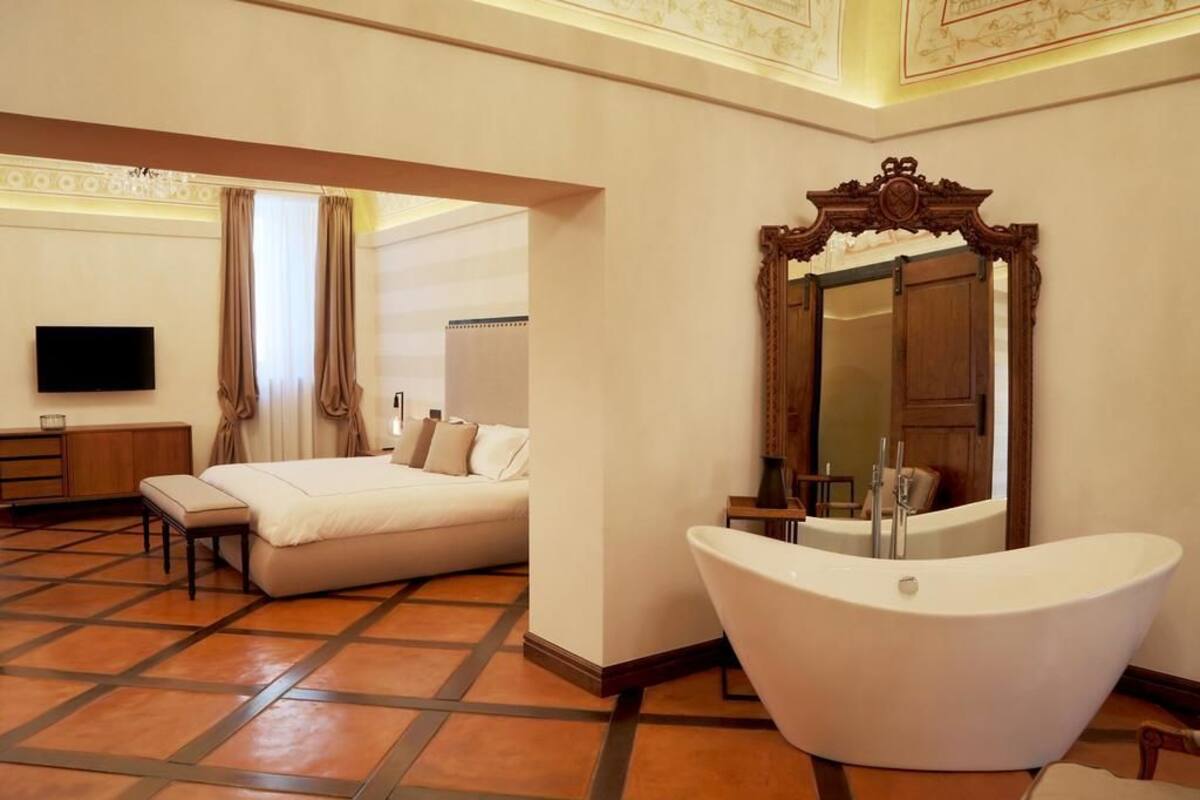 A hotel room with a large bed, a sitting area, a flat-screen TV, a large mirror, and a freestanding bathtub.