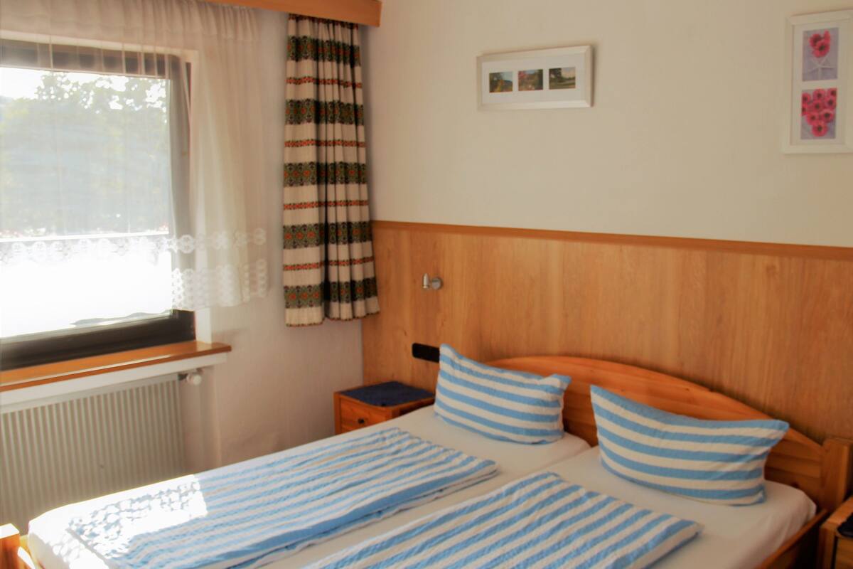 Quarto com cabeceira de madeira, cama com roupa de cama listrada, mesa de cabeceira, janela com cortina translúcida e radiador.