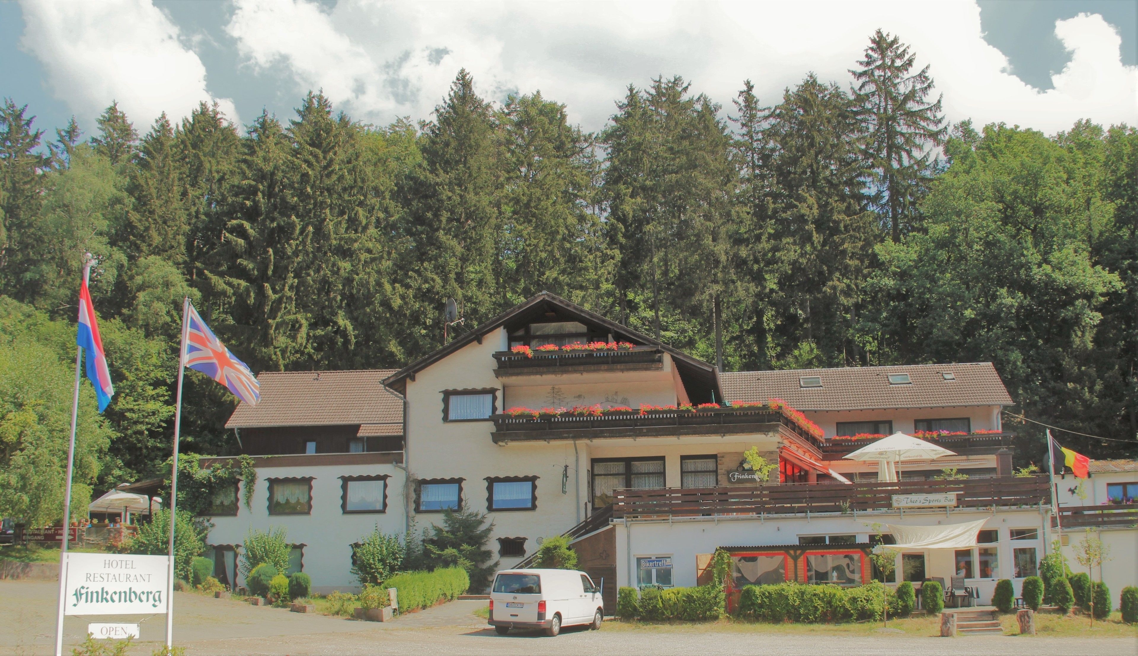A hotel with a balcony, a sign indicating it is open, and a van parked in front.