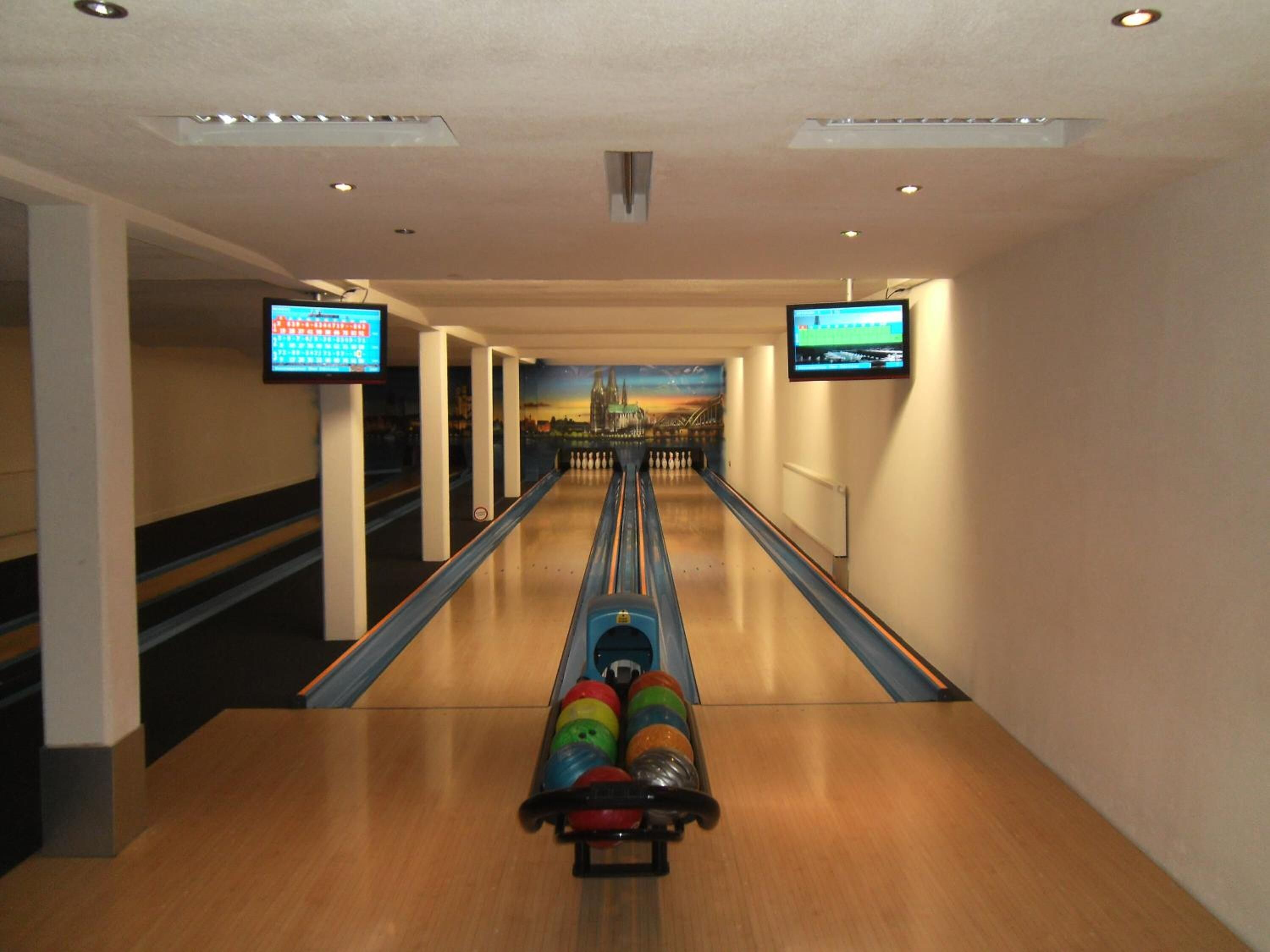 Eine Kegelbahn mit mehreren Bahnen, zwei Bowlingkugeln im Vordergrund und zwei Bildschirmen, die Kegelspiele zeigen.