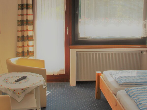 Una habitación con cama, una silla, una mesita con control remoto y una ventana con cortinas.