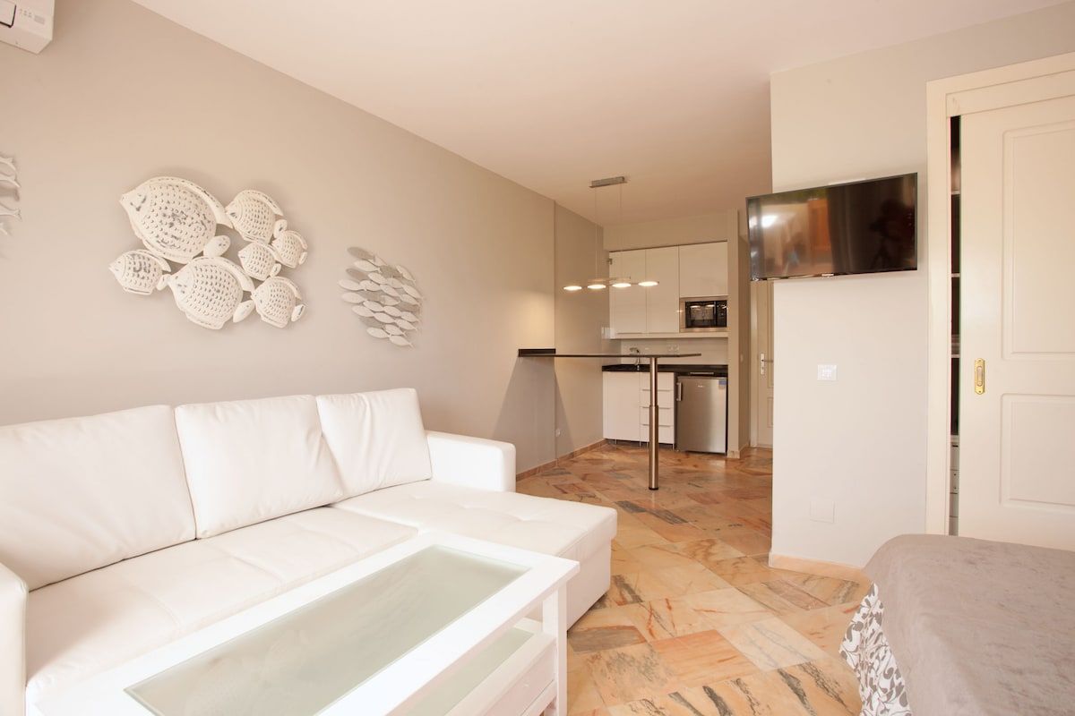 A modern living room with a white sofa, a glass coffee table, and a kitchen in the background.