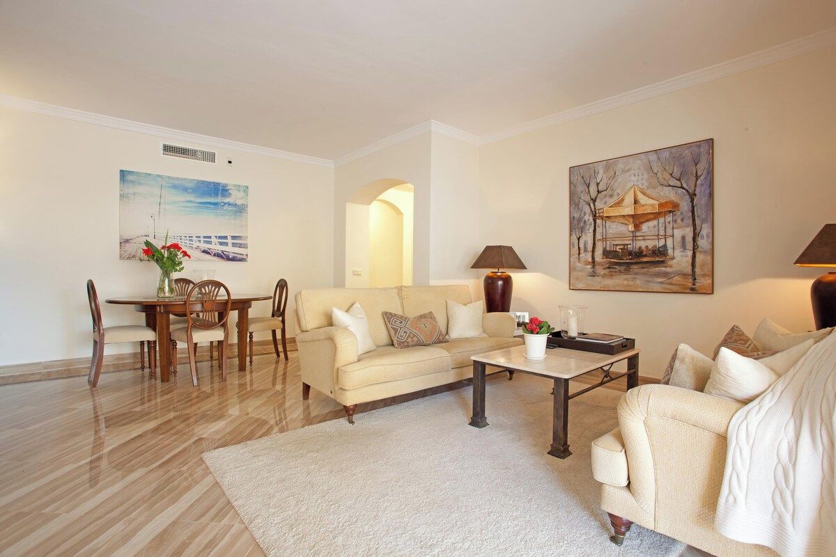 A living room with a sofa, coffee table, and dining area. There are paintings on the walls and a lamp on the table.