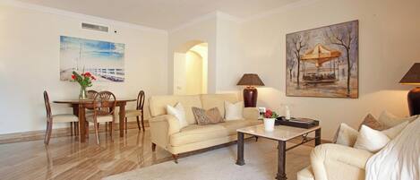 A living room with a sofa, coffee table, and dining area. There are paintings on the walls and a lamp on the table.