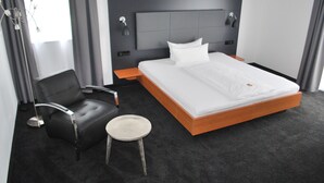 Una habitación de hotel moderna con una cama grande, un sillón de cuero negro, un taburete pequeño y una mesita de noche.