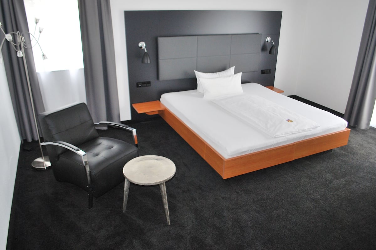 A modern hotel room with a large bed, a black leather chair, a small stool, and a bedside table.