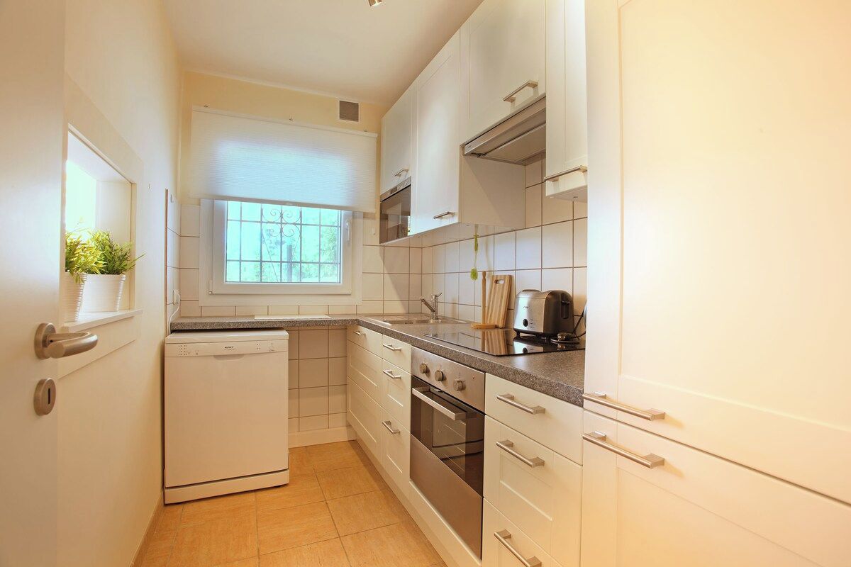 A compact kitchen with white cabinetry, a built-in oven, a dishwasher, and a window providing natural light.