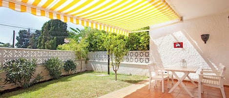 Eine Terrasse mit gestreiftem Sonnensegel, weißen Gartenmöbeln und einem weißen Zaun.