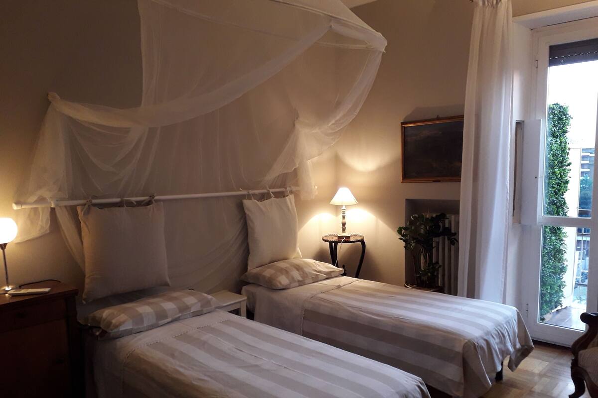 A bedroom with two beds, a bedside table with a lamp, a bedside plant, and a window with a view of greenery.