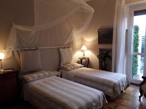 A bedroom with two beds, a bedside table with a lamp, a bedside plant, and a window with a view of greenery. - Light Charme (Milan)