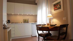 A compact kitchen with white cabinets, a dining table with chairs, a lamp, and a painting on the wall. - Light Charme (Milan)