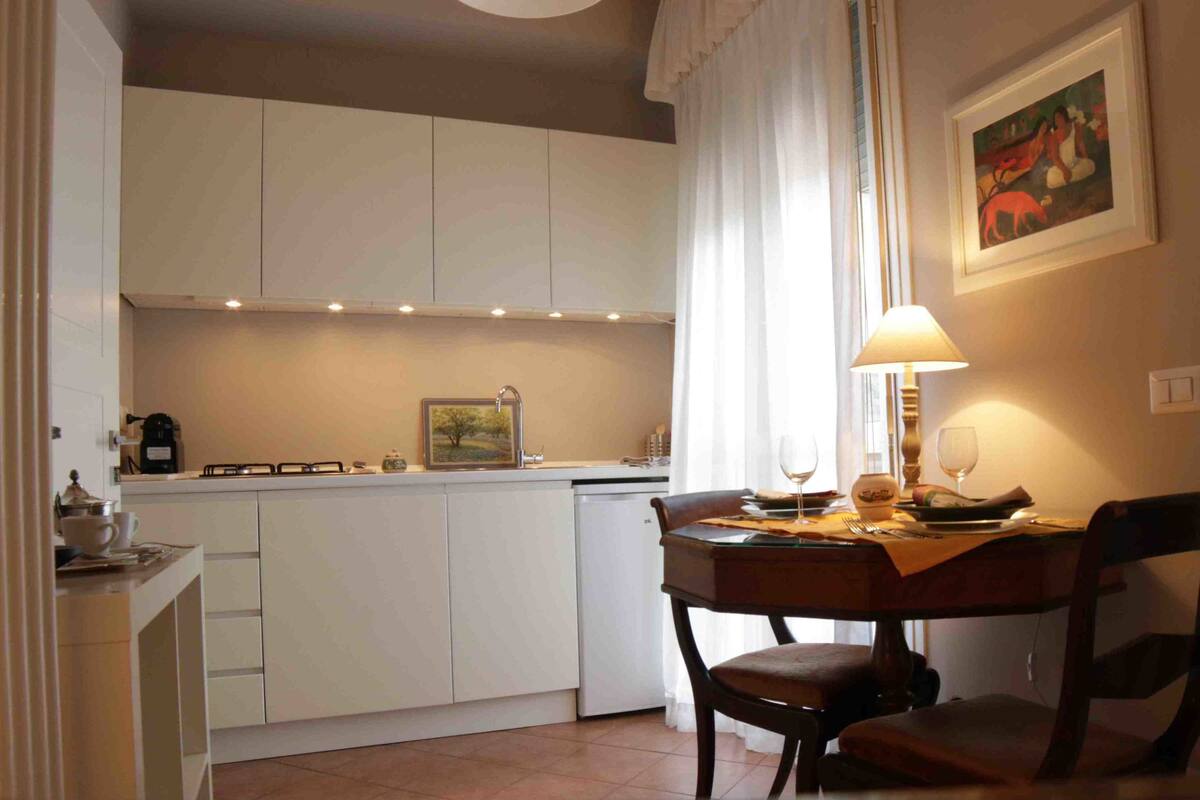 A compact kitchen with white cabinets, a dining table with chairs, a lamp, and a painting on the wall.