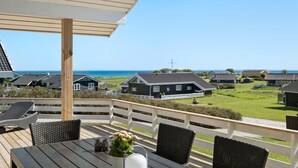 Terraza de madera con mesa y sillas, con vista a una zona costera con casas y el mar.