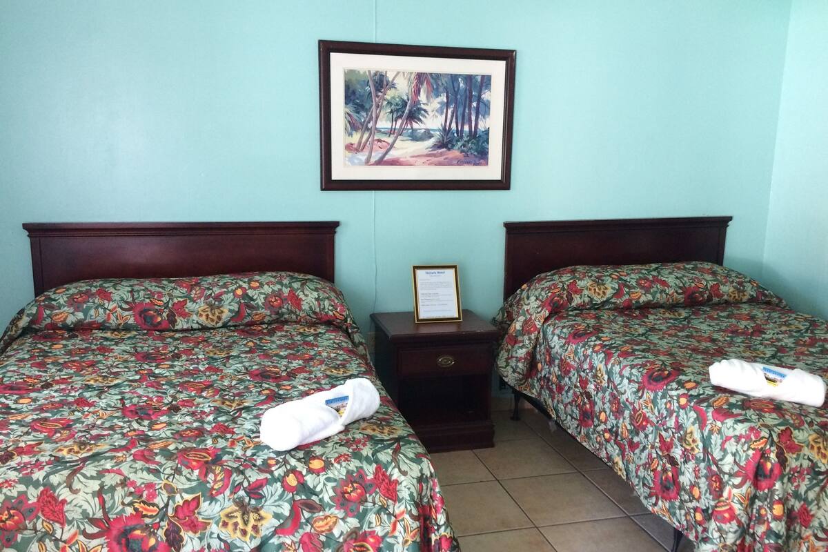 A hotel room with two beds, each with floral bedding, a framed picture on the wall, and a small bedside table with a sign.