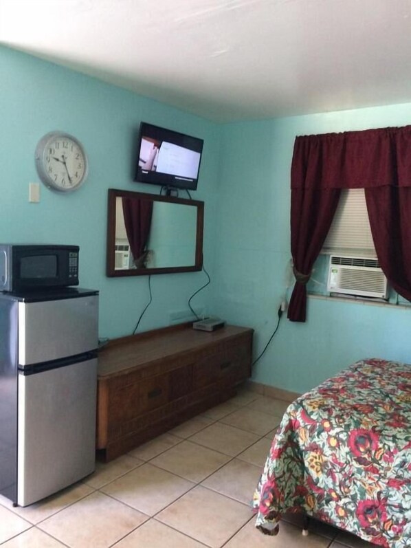 Une pièce aux murs de couleur turquoise, avec une commode en bois, un réfrigérateur, une télévision, une horloge et un lit recouvert d’une couvre-lit à motifs floraux.