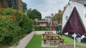 Un espace patio aménagé avec des tables et des bancs en bois, entouré de gazon synthétique verdoyant et d  ’un feuillage dense.