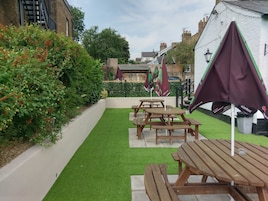 Un área de patio con mesas y bancos de madera, rodeada de césped artificial verde y abundante follaje.