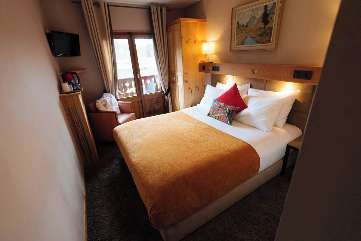 A hotel room with a bed, a wooden wardrobe, a television mounted on the wall, and a window with curtains.