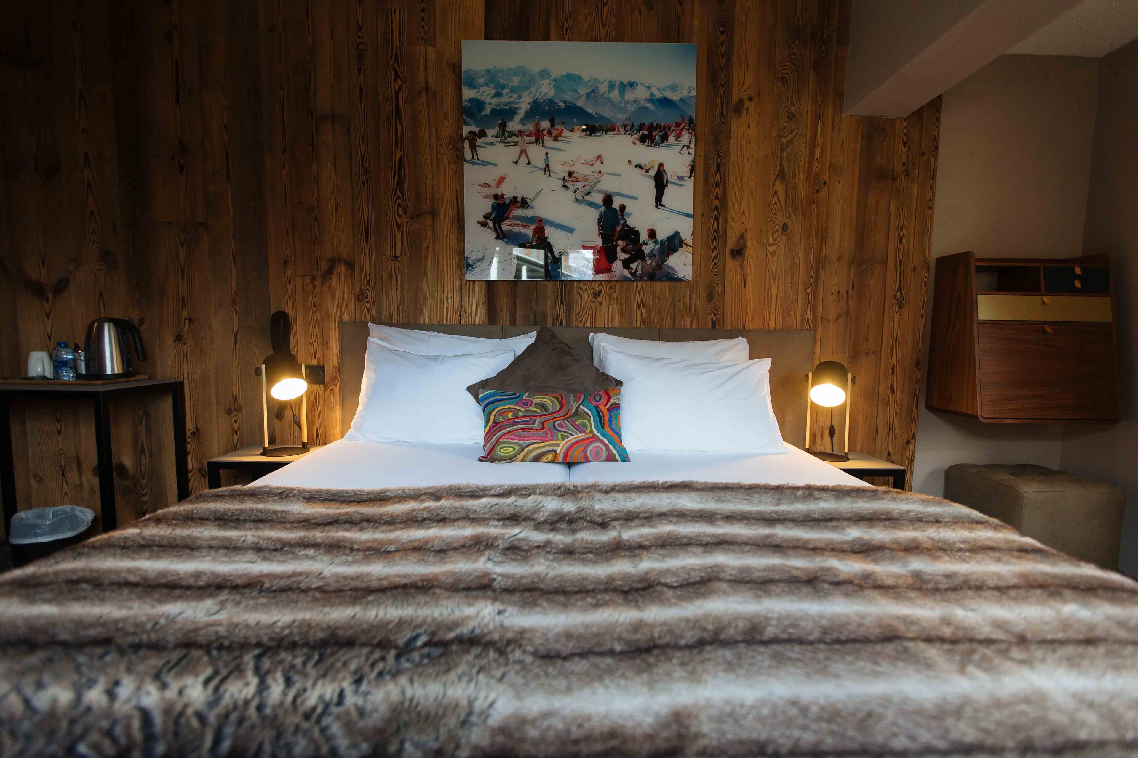 Un lit recouvert d’un plaid en fourrure, des murs en bois, un tableau représentant un paysage enneigé et des lampes de chevet.