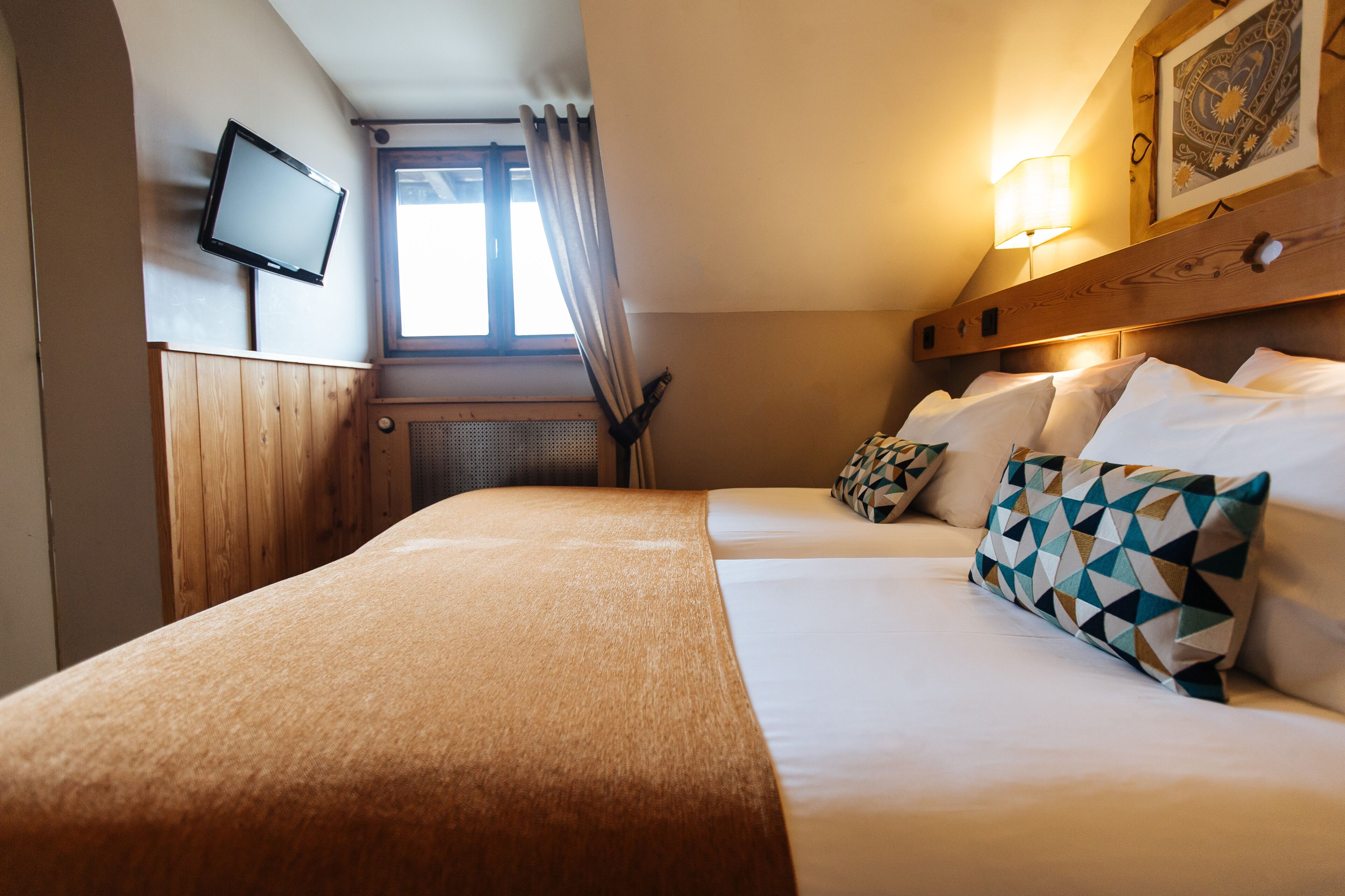 Habitación de hotel con una cama, televisor montado en la pared, ventana con cortinas y una lámpara.