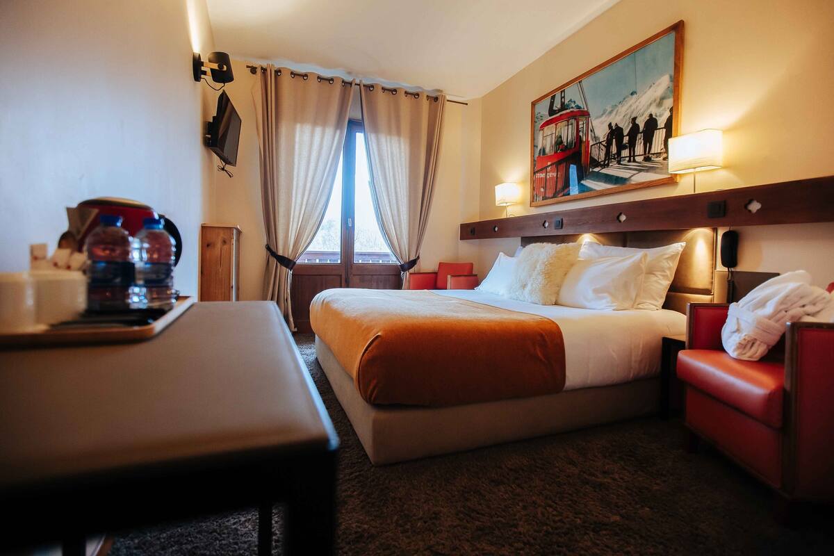 A hotel room with a bed, a desk, a chair, a painting, and a window with curtains.