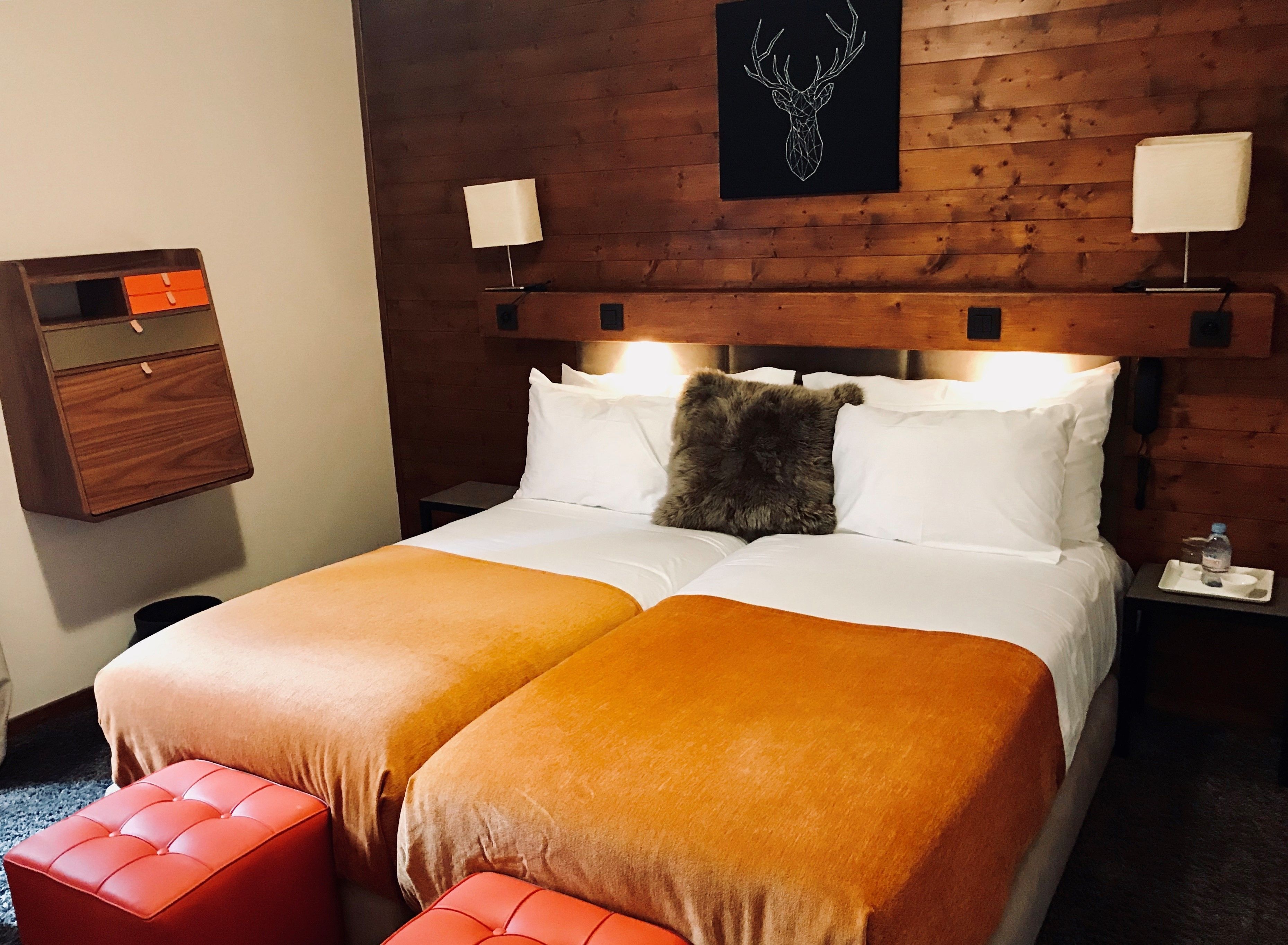 A double bed with two orange blankets, a wooden headboard, a wall-mounted lamp, and a small bedside table with a water bottle.