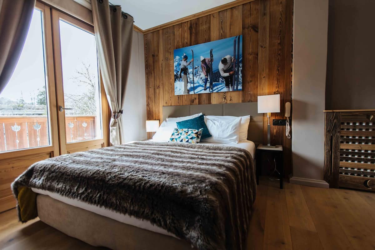 A bedroom with a wooden headboard, a large window with curtains, a painting of skiers, and a fur throw on the bed.