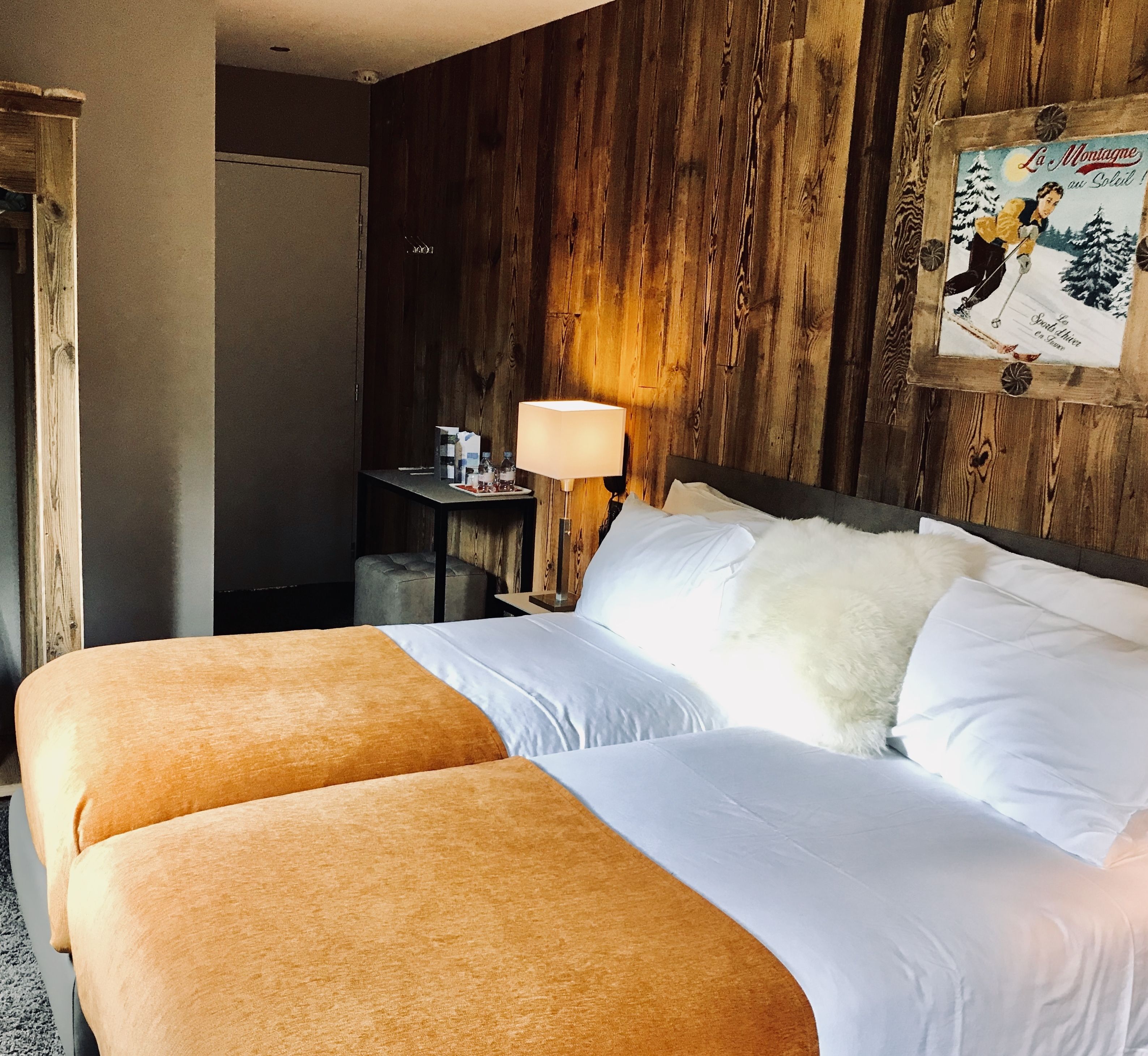 A hotel room with two beds, wooden paneling, a nightstand with a lamp, and a framed picture on the wall.