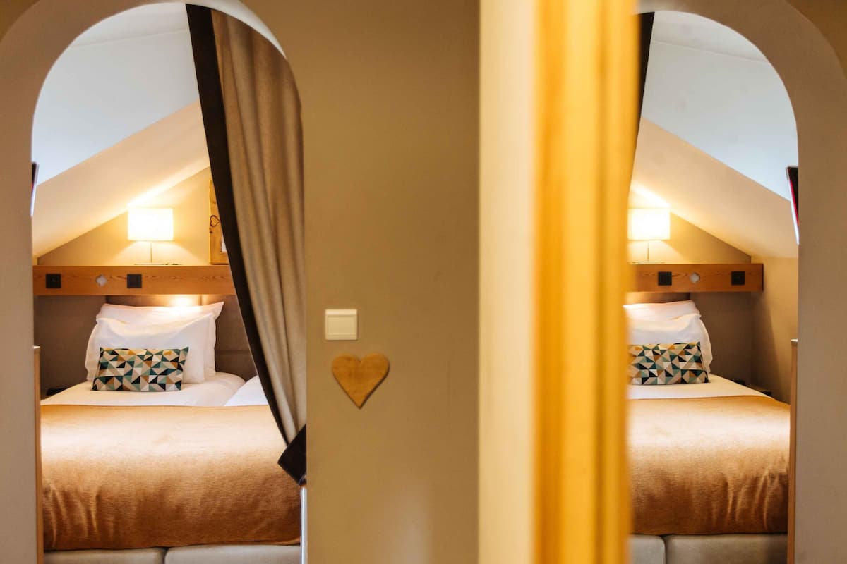 A bedroom with a sloped ceiling, a bed with a geometric-patterned pillow, a heart-shaped decoration on the wall, and a bedside lamp.