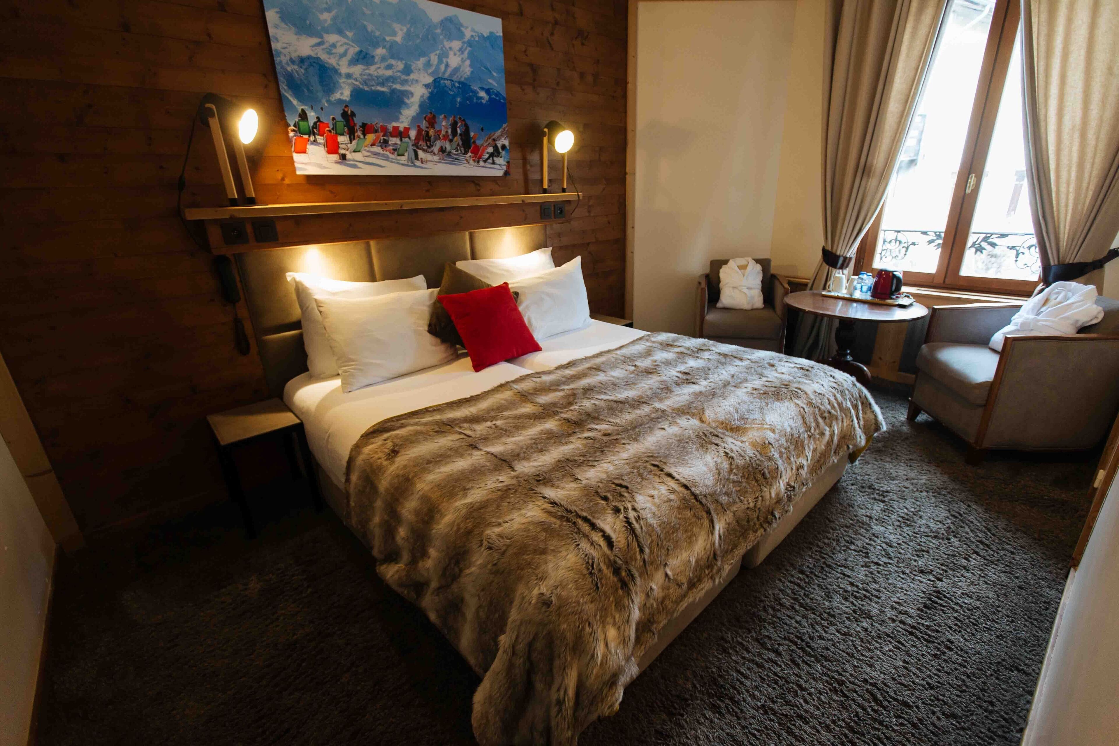 A hotel room with a large bed, a headboard with lights, a small table with a chair, and a window with curtains.