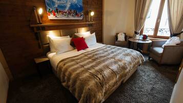 A hotel room with a large bed, a headboard with lights, a small table with a chair, and a window with curtains.